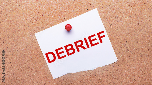White paper with the text DEBRIEF is attached to the wooden background with a red button.