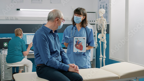 Принти на полотні Female assistant holding tablet with cardiovascular system illustration, explaining heart condition to retired person during covid 19 pandemic