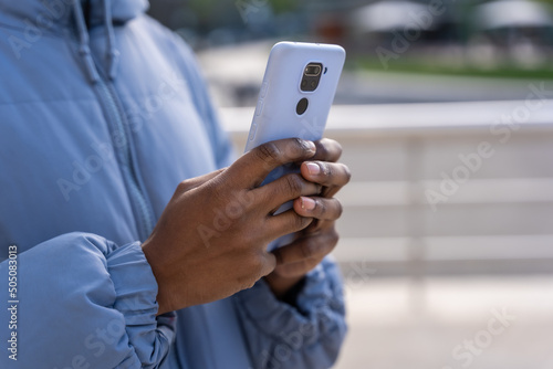 Hands of unrecognizable black ethnic man with phone and blue jacket in the city
