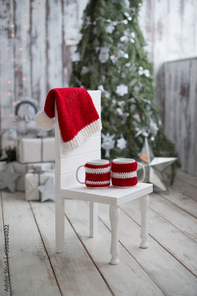 Obraz premium Two mugs in knitted covers with a Santa hat are lying on a white wooden chair, Christmas background.