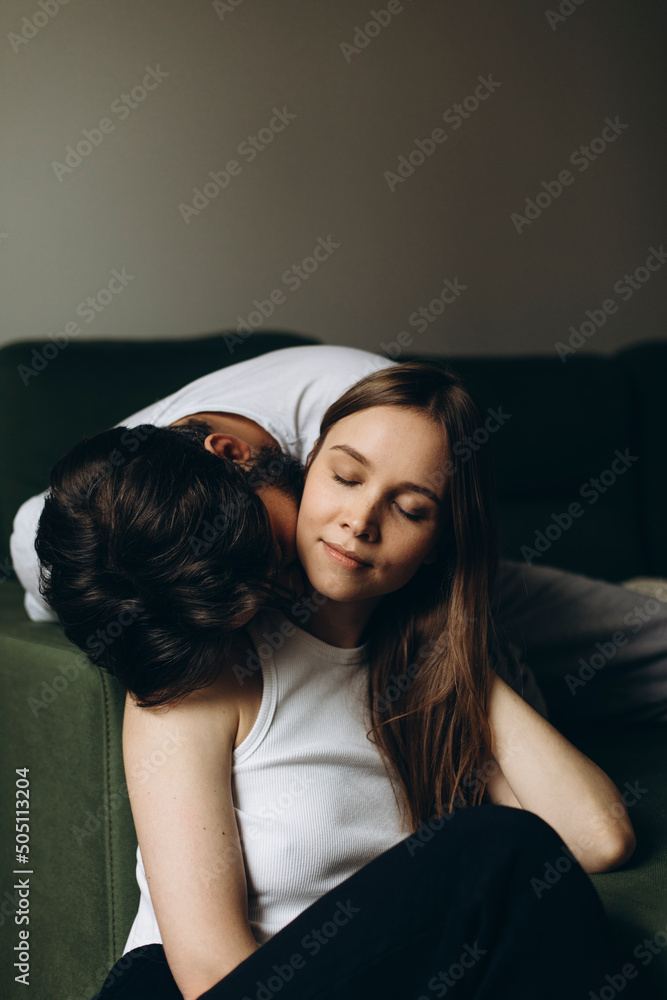 a dark-haired young man tenderly kisses the neck of a blonde-haired young woman indoors. young loving couple. beautiful woman closed her eyes from enjoying the kiss a dark-haired young man tenderly kisses the neck of a blonde-haired young woman indoors. young loving couple. beautiful woman closed her eyes from enjoying the kiss