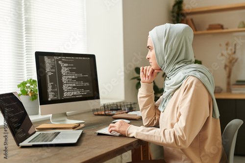 Successful Muslim programmer wearing hijab sitting at desk working on new software using desktop PC and laptop, copy space