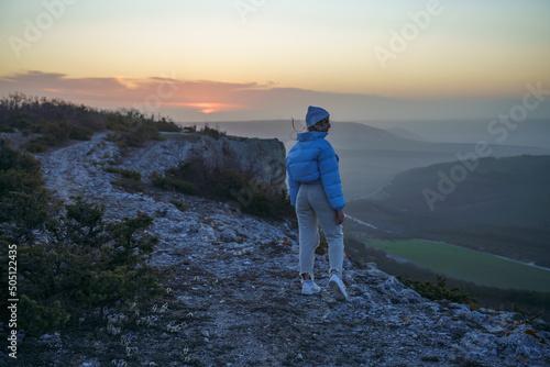 Woman tourist on top of sunrise mountain. The girl salutes the sun, wearing a blue jacket, white hat and white jeans. Conceptual design.