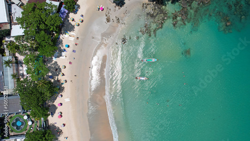 The snow-white Kata beach of Phuket island. Top view from a drone on the ocean waves. There are several Thai boats. The whole island is green. The water is clear and transparent. People are bathing
