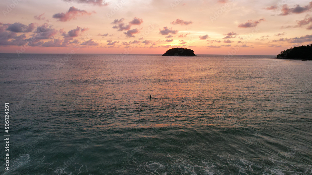 A surfer girl is waiting for a wave at sunset. It is located alone in the sea. Admires the view of the island, the orange-red sky and clouds. All this is reflected in the sea. Extreme romance. Epic A surfer girl is waiting for a wave at sunset. It is located alone in the sea. Admires the view of the island, the orange-red sky and clouds. All this is reflected in the sea. Extreme romance. Epic