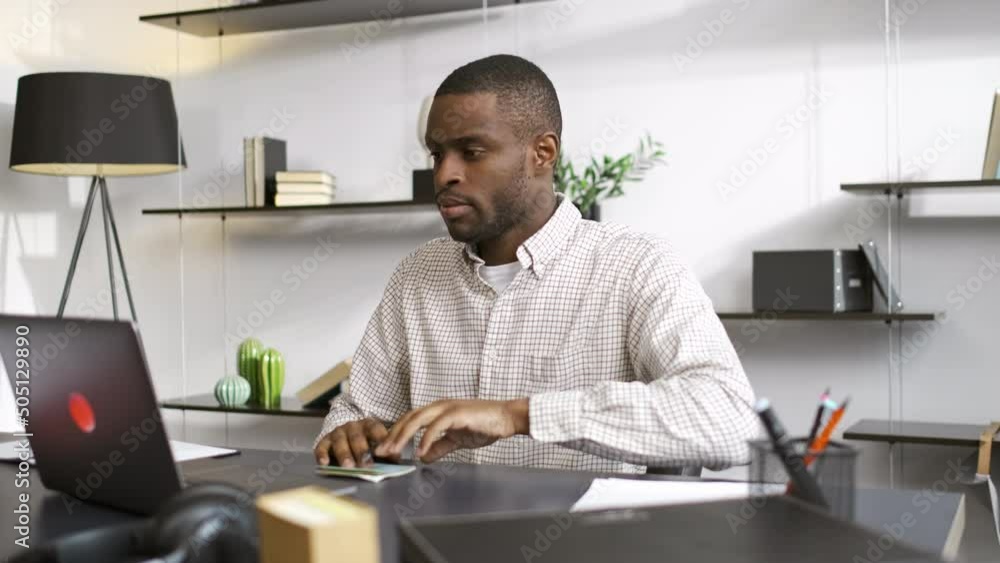 Young man is looking at laptop and writing while sitting at table in home office msspe. 4k Close view of bearded african american guy looks at device display and takes notes on paper, plans work day Young man is looking at laptop and writing while sitting at table in home office msspe. 4k Close view of bearded african american guy looks at device display and takes notes on paper, plans work day