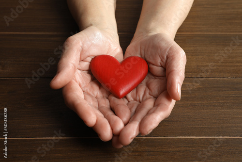 Elderly woman holding red heart in hands at wooden table, closeup
