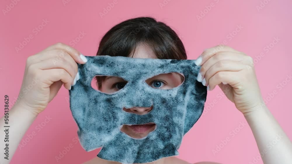 Portrait of a young woman who is holding sheet bubble mask close-up on a pink background. Portrait of a young woman who is holding sheet bubble mask close-up on a pink background.
