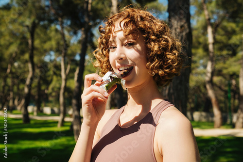 Eating protein bar, redhead woman wearing sport bra eating protein bar while standing on a city park, outdoors.Resting after sports training. Nutrition for sport. Healthy muesli and cereal snacks.