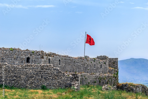 The red Albanian flag rises above the ruins of the walls of the ancient Shkoder castle in Albania. The remains of an ancient fortress among the green grass against the blue sky photo