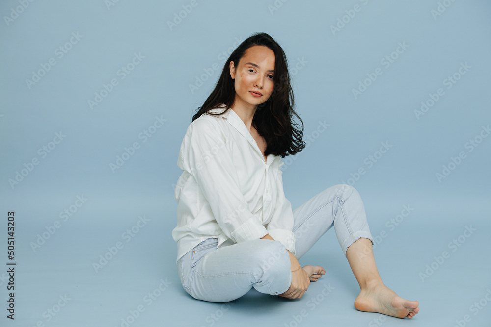 Casual beautiful vitiligo brunette sitting barefoot, presenting deseased patchy part of her face over blue background. Looking peacefully at the camera. Casual beautiful vitiligo brunette sitting barefoot, presenting deseased patchy part of her face over blue background. Looking peacefully at the camera.