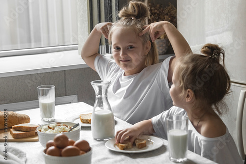 two sister girls in white t-shirts have breakfast at home in the kitchen with natural and healthy products  cottage cheese and cornbread  milk and boiled chicken eggs