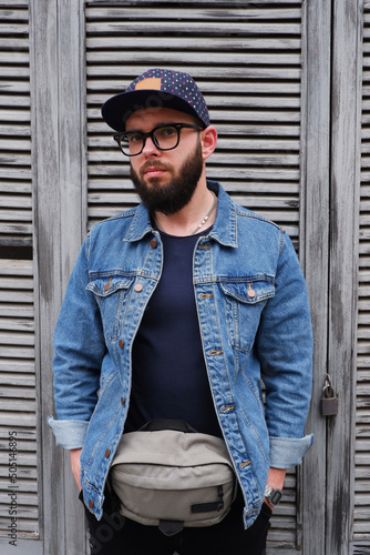 Handsome hipster guy in a cap and glasses with a beard in a blue denim jacket and a fanny pack.