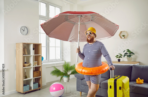 Plus size man in striped swimsuit, hat, shades and floaty standing in living room, holding beach umbrella and luggage and making funny face. Covid 19 pandemic, summer holiday, vacation at home concept