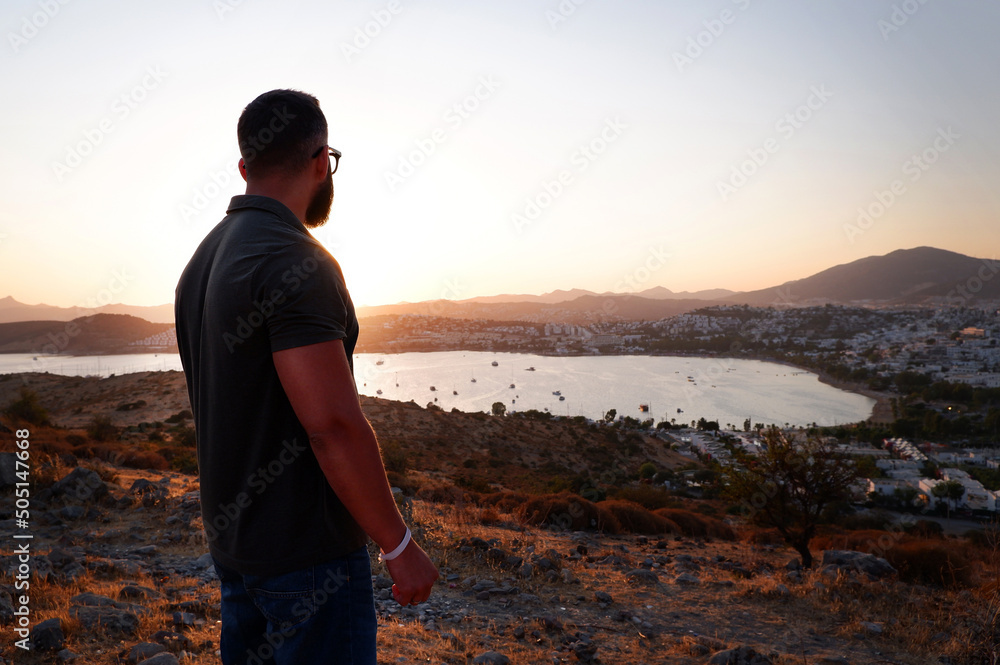 A young buff tourist with a beard and glasses in a dark T-shirt stands on a mountain with his back to the camera overlooking the bay