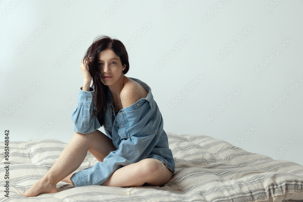 Portrait of young beautiful woman sitting on mattress, posing in big male shirt isolated over grey studio background Portrait of young beautiful woman sitting on mattress, posing in big male shirt isolated over grey studio background