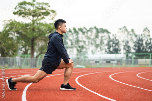 Young asian man wearing sportswear warming up front of sport stadium. Chinese man stretching legs before running on track exercise in the morning. Healthy and athlete lifestyle concept.