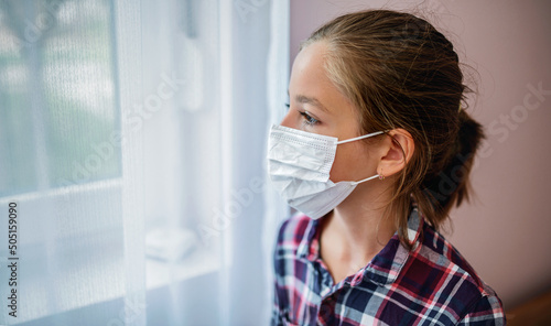 Quarantine in corona time. Worried little girl with medical mask on her face watching through the window. Medical concept