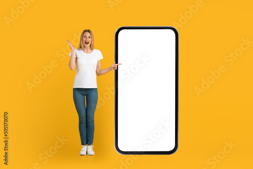 Glad shocked young european woman with open mouth in white t-shirt points finger at huge phone with blank screen
