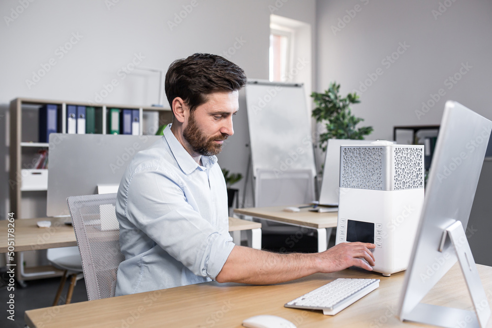 Young handsome man, office worker, works includes humidifier, air conditioner which stands on the table near the computer in the office Young handsome man, office worker, works includes humidifier, air conditioner which stands on the table near the computer in the office