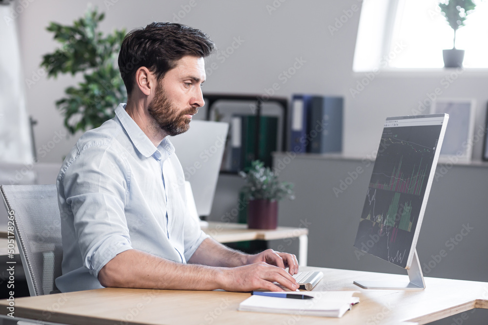 Young handsome man, office worker, working at a computer with graphics, sitting at a table, typing on the keyboard, explains