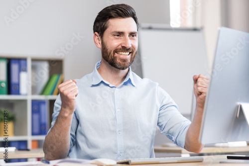 Young successful man, office worker, manager, freelancer rejoices in the result of the work done. He sits happy at the table and computer, rejoices, raises his hands