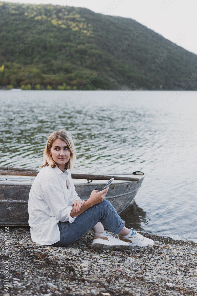 young woman in white shirt and jeans with a phone in her hands sits on boat on the shore of mountain lake