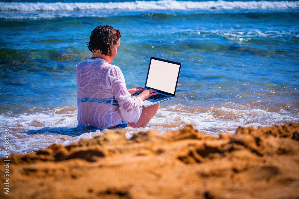 Young beautiful woman working remotely, using laptop computer by the sea. Freelance work concept. Horizontal image.