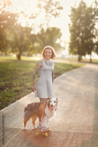 Cute hipster woman girl with tattoo rides skateboard with an Australian Shepherd dog on sidewalk in park, warm summer evening, training dog