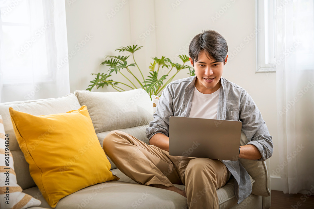 Asian freelance man sitting on sofa at home working on laptop in the living room, Technology and lifestyle concept. 