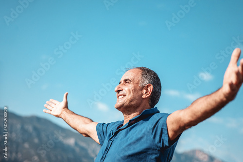 An aged happy smiling person standing with outstretched arms on mountains and blue sky background.