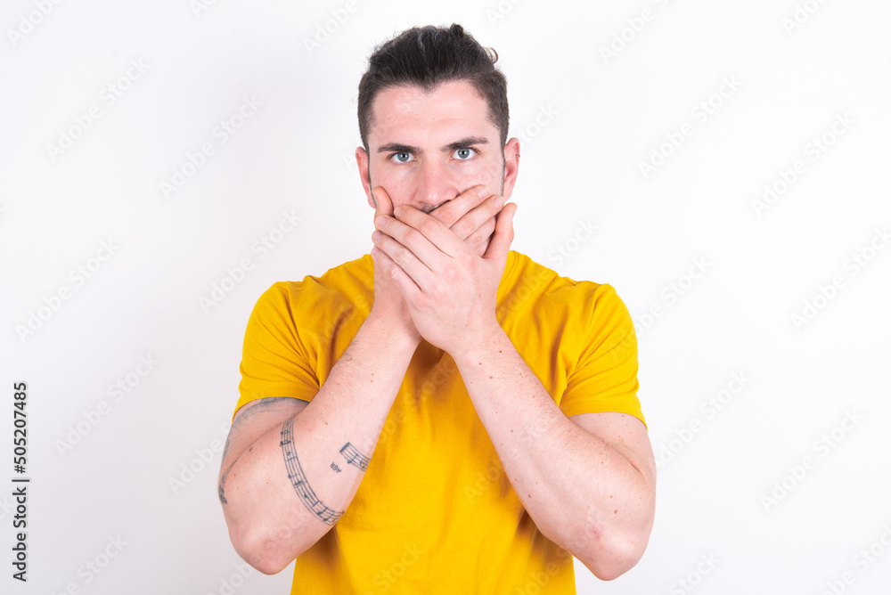 Stunned young dark haired man wearing yellow t-shirt over white background covers both hands on mouth, afraids of something astonishing Stunned young dark haired man wearing yellow t-shirt over white background covers both hands on mouth, afraids of something astonishing