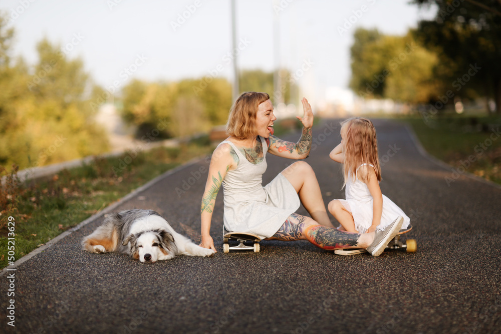 Stylish crazy European hipster mom with tattoos with her daughter child and dog Australian Shepherd breed. On skateboards and pavement in park, warm summer evening and family time and hobbies Stylish crazy European hipster mom with tattoos with her daughter child and dog Australian Shepherd breed. On skateboards and pavement in park, warm summer evening and family time and hobbies