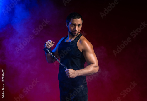 muscular man with a chain. sportsman posing in black t-shirt, jeans. studio portrait. red background blue smoke around the guy