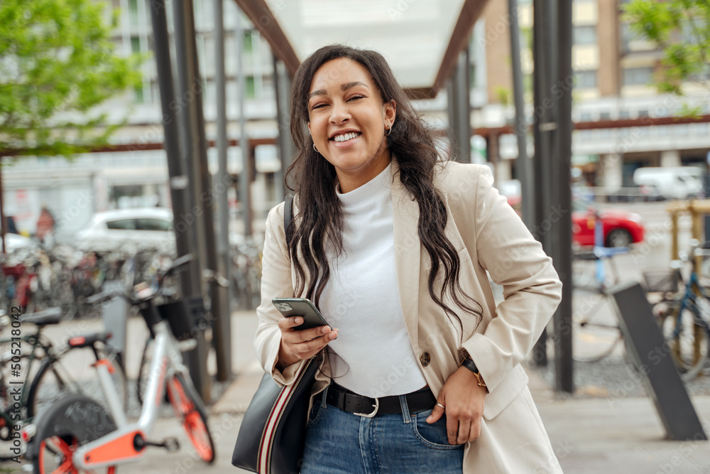 Beautiful confident woman with emotional face holding mobile phone looking at camera, laughing on the street 