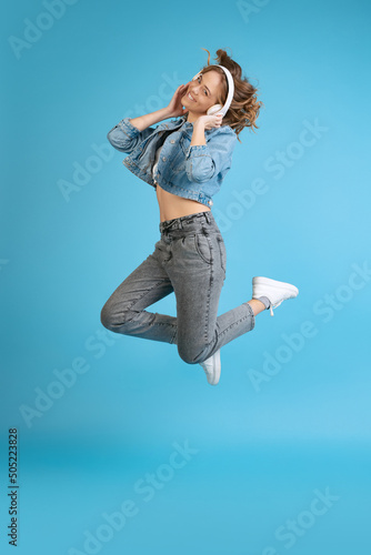 Portrait of young excited girl listening to music in headphones isolated on blue background. Concept of beauty, music, fashion, youth, spring collection