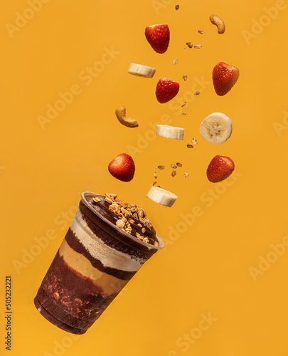 Acai cup, layers of granola, ground peanuts, and pieces of fruits and nuts flying in the air, yellow background.
