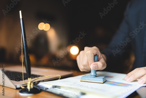 Close-up of a person's hand stamping with approved stamp on certificate document public paper at desk, notary or business people work from home, isolated for coronavirus COVID-19 protection photo