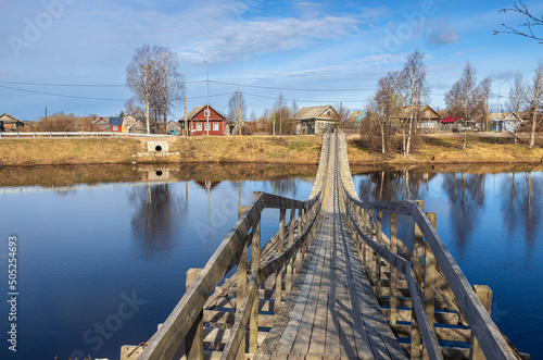 Verkhovye, Olonetsky district, Karelia, Russia, - April 28, 2022,, Narrow wooden pedestrian suspension bridge across the river in russian village near Olonets. Sunny summer evening.