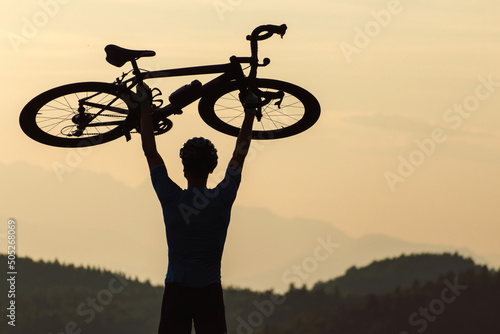 Male cyclist silhouette celebrating a good race, raising a bicycle above the head and looking at the beautiful sunset over mountain landscape