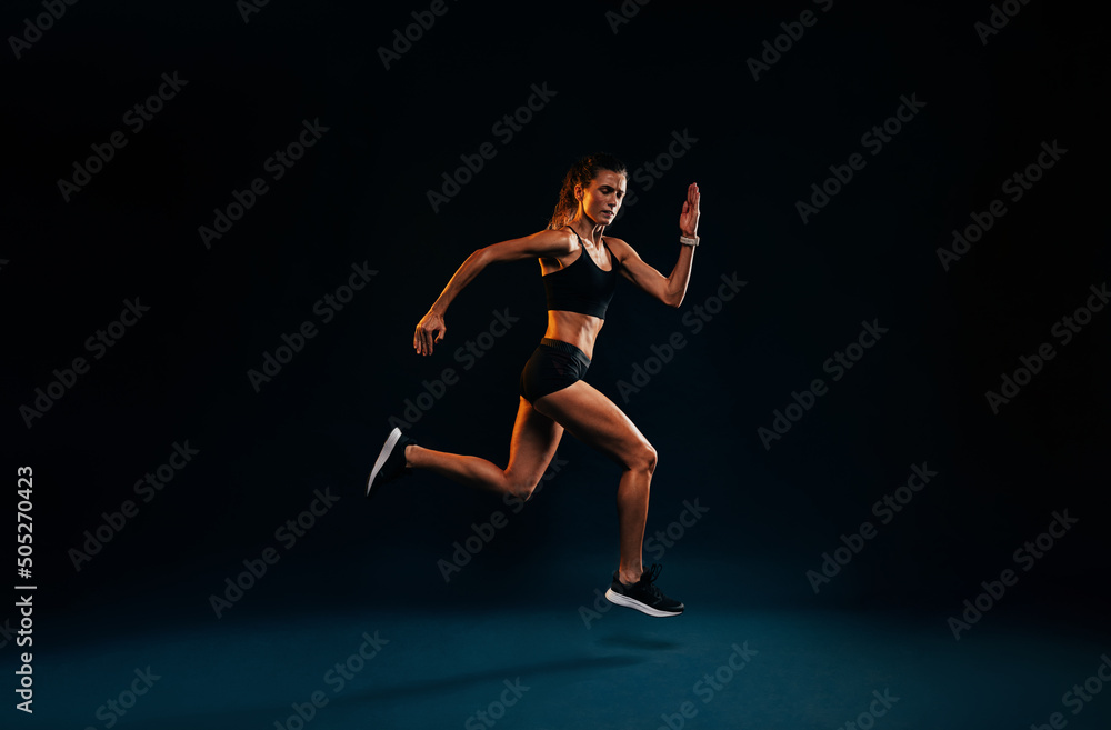 Female runner sprinting on black background. Fit woman jumping while exercising in studio.