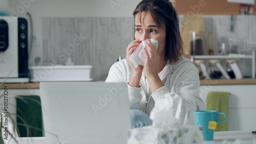 Video of llness young woman sneezing in a tissue while working with laptop sitting in the kitchen at home.
