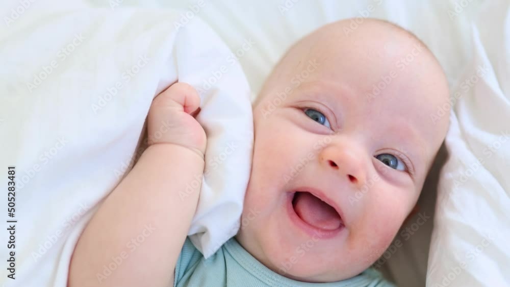 funny infant face portrait, blue eyes happy smiling baby child 3 months on white sheets. caucasian kid close-up portrait. happy little baby lying in bed looking into camera making funny grimaces. funny infant face portrait, blue eyes happy smiling baby child 3 months on white sheets. caucasian kid close-up portrait. happy little baby lying in bed looking into camera making funny grimaces.