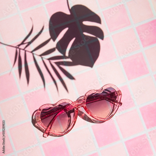 Sunglasses, palm and monstera leaf shadow on pink mosaic tiles. Minimalistic summer concept. Sunny day composition.