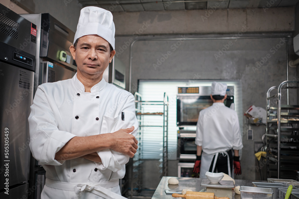 Professional senior Asian male chef in white cooking uniform, food occupation looks at camera, arms crossed, expertise in commercial pastry culinary jobs, catering restaurant kitchen.