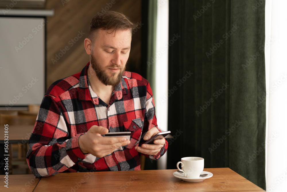 bearded young man holding a mobile phone and a credit card. businessman checking account balance, using online banking app on his mobile phone, drinking coffee in cafe bearded young man holding a mobile phone and a credit card. businessman checking account balance, using online banking app on his mobile phone, drinking coffee in cafe