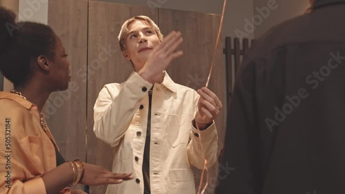 Medium slowmo of young gay man with dyed blonde hair holding balloon on string while talking to multiethnic LGBTQ friends, gathering together for small dinner party photo