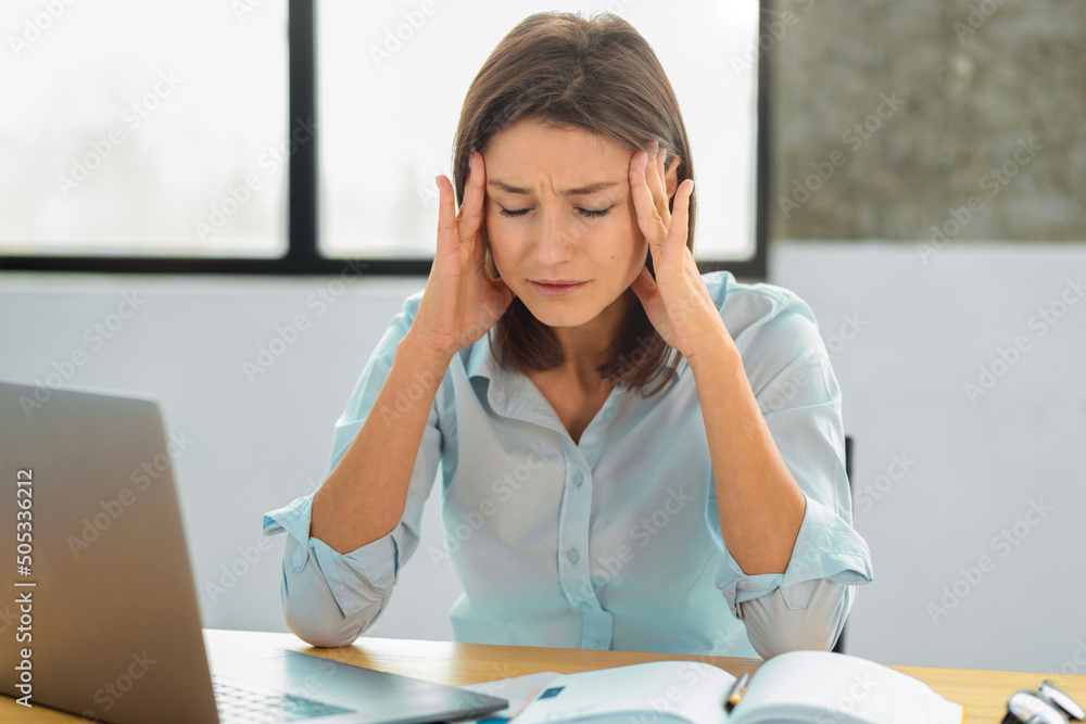 Portrait of a young caucasian woman employee with headache rubbing her temples sitting in the office. Overworked female concept Portrait of a young caucasian woman employee with headache rubbing her temples sitting in the office. Overworked female concept