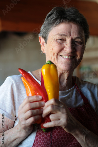 Happy portrait of an older housewife wearing her homemade apron and holding three different colored palermo peppers in her hands.