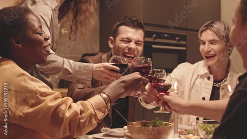 Medium slowmo of young diverse LGBTQ friends in fancy clothes cheering with glasses of red wine while having cozy dinner party at festively decorated table with candles photo